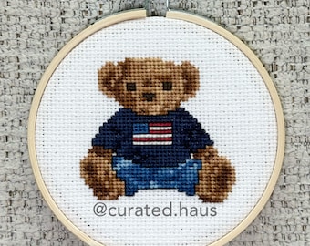 Peut inclure: Une broderie au point de croix dans un tambour en bois circulaire représentant un ours en peluche marron portant un t-shirt bleu foncé avec un motif de drapeau américain rouge et blanc. Le texte @curated.haus est brodé en gris foncé.