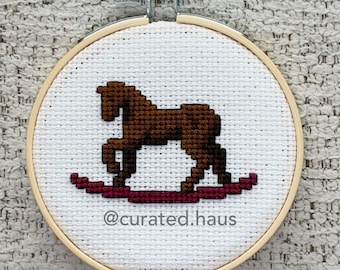 Peut inclure: Un tambour à broder rond en bois contenant une toile Aida blanche avec un motif au point de croix représentant un cheval à bascule marron sur une base rouge foncé. Le texte @curated.haus est brodé en gris foncé sous le cheval.