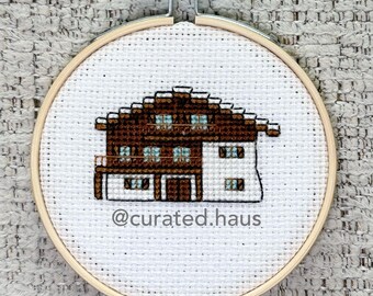 Peut inclure: Une broderie au point de croix dans un tambour en bois circulaire représentant un chalet marron et blanc avec des fenêtres bleues. Le tissu blanc affiche le texte @curated.haus en police sans-serif gris foncé sous la maison.