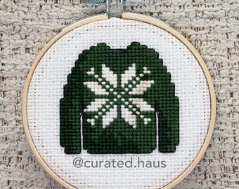 Peut inclure: Une broderie au point de croix dans un tambour en bois circulaire représentant un pull vert foncé avec un motif de flocon de neige blanc sur le devant. Le texte @curated.haus est brodé en fil gris foncé sous le pull sur le tissu blanc.