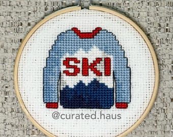 Peut inclure: Une broderie au point de croix dans un tambour en bois circulaire représentant un pull bleu clair et bleu foncé avec le mot SKI en lettres rouges. Le texte @curated.haus est brodé en gris au bas du tissu blanc en arrière-plan.