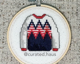 Peut inclure: Une broderie au point de croix dans un tambour en bois circulaire représentant un pull avec un motif géométrique rouge, blanc et bleu marine. Le texte @curated.haus est brodé en fil gris sous le pull sur le tissu blanc.