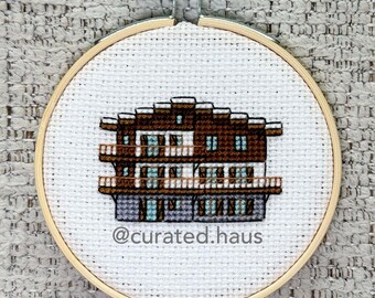 Peut inclure: Une broderie au point de croix représentant un chalet à plusieurs étages avec du fil marron, blanc et bleu clair, encadrée dans un tambour en bois circulaire. Le texte @curated.haus est brodé en gris foncé sous le bâtiment sur le tissu blanc.