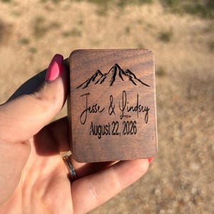 May include: A rectangular dark brown wooden box featuring a laser-engraved mountain range graphic at the top, followed by the names Jesse & Lindsey in a cursive script, and the date August 22, 2026, printed in a clean sans-serif font.
