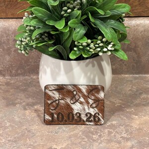 May include: A rectangular patch featuring brown and white cowhide texture with the engraved letters J & S and the date 10.03.26. The patch is positioned in front of a white ceramic pot containing a lush green plant with small white berries.