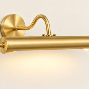 May include: A wall-mounted picture light with a brushed brass finish. The fixture features a circular base plate, a curved arm, and a horizontal cylindrical lamp head with decorative end caps. A warm light emits from the bottom of the cylinder.