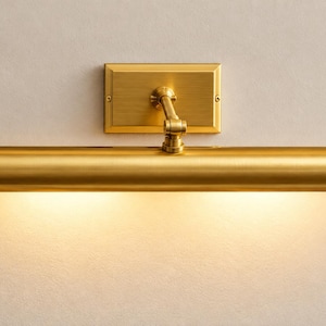 May include: A wall-mounted picture light with a brushed brass finish. The fixture features a rectangular backplate, an adjustable articulated arm, and a long cylindrical tube housing the light source, which emits a warm glow downward.