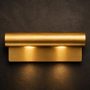 May include: A horizontal wall sconce with a brushed brass finish, featuring a cylindrical top bar and a rectangular backplate. The fixture emits a warm, focused light downward from beneath the cylindrical element, creating a soft glow.