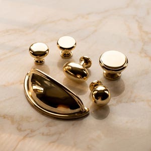 May include: A collection of polished gold-toned hardware including one large semi-circular cup pull, one oval-shaped knob, and four round knobs of varying sizes. Each piece features a smooth, reflective metallic finish and a sturdy base.