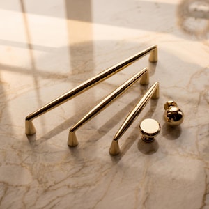May include: A collection of gold-toned hardware including three elongated cabinet pulls of varying lengths with tapered ends, one flat circular knob, and one spherical knob, all featuring a smooth, reflective metallic finish.