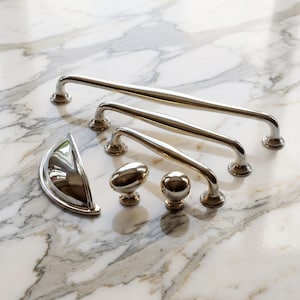 May include: A collection of polished nickel cabinet hardware including three varying lengths of arched bar pulls, one semi-circular cup pull, and two round knobs. Each piece features a smooth, reflective metallic finish and a sturdy construction.
