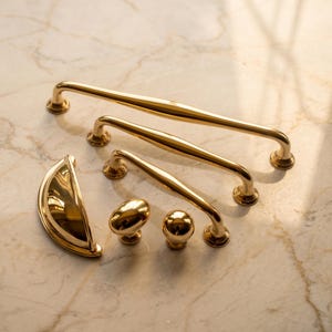May include: A collection of polished gold-toned hardware including three varying lengths of arched cabinet pulls, one semi-circular cup pull, one oval-shaped knob, and one spherical knob, all featuring a smooth, reflective metallic finish.