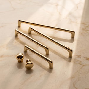 May include: A collection of gold-toned cabinet hardware including three elongated metal drawer pulls with curved ends and two spherical knobs. The hardware features a smooth, reflective metallic finish and a simple, clean-lined structural design.