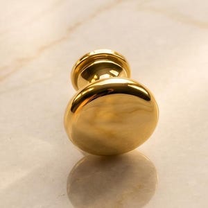 May include: A polished gold-tone cabinet knob with a smooth, rounded, mushroom-shaped head and a cylindrical base. The hardware features a highly reflective, metallic finish that catches light, creating bright highlights across its curved surface.