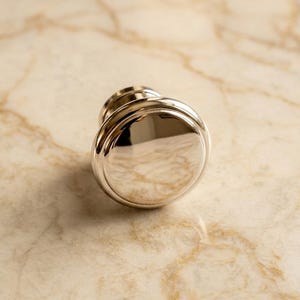 May include: A polished silver-toned metal cabinet knob featuring a smooth, circular face with a raised outer rim and a stepped base. The reflective surface shows a high-gloss finish, highlighting the rounded, convex shape of this hardware piece.
