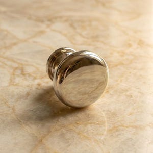 May include: A polished silver-toned metal cabinet knob with a smooth, rounded, mushroom-shaped head and a cylindrical base. The surface of the knob is highly reflective, showing light highlights and a metallic finish throughout the object.