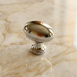 May include: A polished chrome cabinet knob featuring a smooth, rounded oval head and a cylindrical base. The metallic surface is highly reflective, showing distorted light reflections of the surrounding environment on its curved exterior.