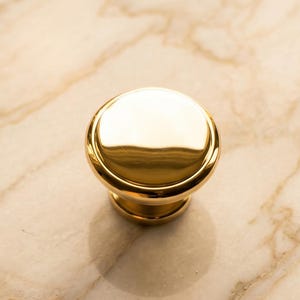 May include: A polished gold-tone metal cabinet knob featuring a smooth, rounded top surface and a tiered circular base. The hardware has a reflective, high-gloss finish that catches light, creating a bright, metallic appearance on the rounded head.