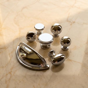 May include: A collection of polished chrome cabinet hardware including one semi-circular cup pull and six assorted knobs in spherical, flat-topped, and oval shapes. Each piece features a reflective, smooth metallic finish with a bright silver tone.
