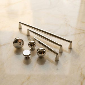 May include: A collection of polished silver-toned hardware including three elongated cabinet pulls of varying lengths and three round cabinet knobs with a smooth, reflective metallic finish and a simple, clean design for furniture or cabinetry.