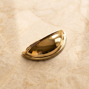 May include: A polished gold-tone metal cup pull handle with a smooth, semi-circular design. The hardware features a reflective, high-gloss finish with a curved, recessed grip area and two visible screw holes on the flat mounting flanges.