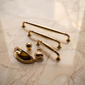 May include: A collection of polished gold-tone cabinet hardware including three varying lengths of arched bar pulls, one round knob, one oval knob, and one semi-circular cup pull, all featuring a smooth, reflective metallic finish.