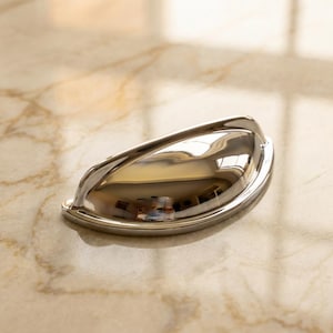 May include: A polished chrome cup pull handle with a smooth, semi-circular design. The hardware features a reflective, mirror-like finish with a curved grip area and two mounting points on the flat base for attachment to cabinetry or furniture.