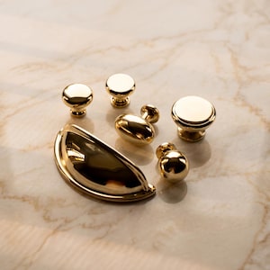 May include: A collection of polished gold-toned hardware including one large semi-circular cup pull, one oval-shaped knob, and four round knobs of varying sizes. Each piece features a smooth, reflective metallic finish and a sturdy base.