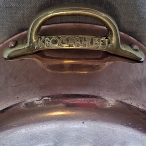 Könnte beinhalten: Nahaufnahme eines Kupfertopfes mit einem Messinggriff. Der Griff ist gewölbt und mit Nieten am Topf befestigt. Der Text "KROGARHUSET" ist auf dem Griff eingraviert. Der Topf hat eine glänzende, reflektierende Oberfläche.