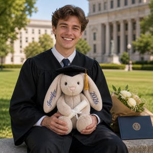May include: A plush bunny wearing a graduation cap and holding a diploma. The bunny has the name "Jennifer" embroidered on one ear and "2026" on the other. The bunny is beige with a black cap and gold tassel. A bouquet of white flowers and a diploma are also present.