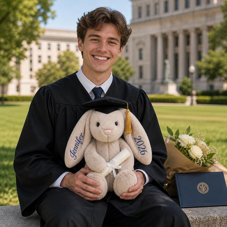 May include: A plush bunny wearing a graduation cap and holding a diploma. The bunny has the name "Jennifer" embroidered on one ear and "2026" on the other. The bunny is beige with a black cap and gold tassel. A bouquet of white flowers and a diploma are also present.