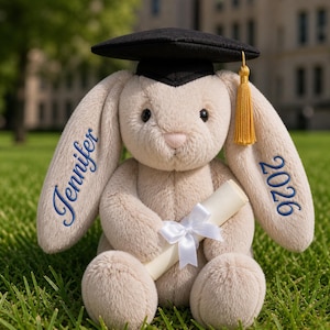 May include: A plush bunny wearing a black graduation cap with a gold tassel. The bunny holds a rolled-up scroll tied with a white ribbon. The name "Jennifer" is embroidered in blue on one ear, and "2026" on the other. The bunny is sitting on green grass.