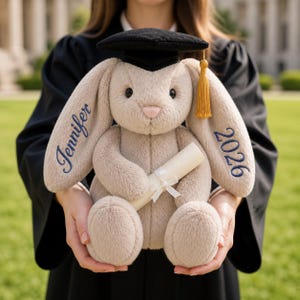 May include: A plush beige bunny wearing a black graduation cap with a gold tassel. The bunny holds a rolled-up diploma tied with a white ribbon. The name "Jennifer" and the year "2026" are embroidered in blue on the bunny's ears. A graduation gift.