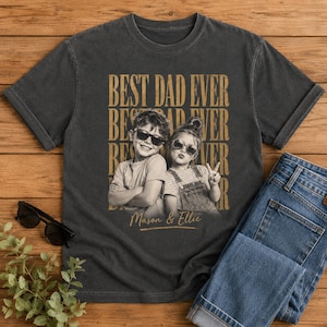 May include: Dark gray t-shirt with the text "BEST DAD EVER" in a stacked design. The shirt features a black and white photo of two children wearing sunglasses. The names "Mason & Ellie" are printed below the photo. Blue jeans are folded next to the shirt.