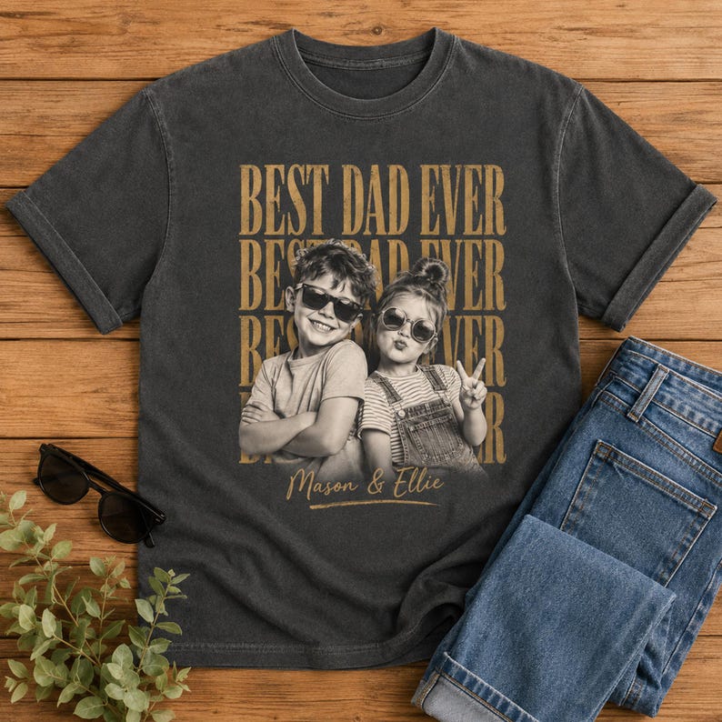 May include: Dark gray t-shirt with the text "BEST DAD EVER" in a stacked design. The shirt features a black and white photo of two children wearing sunglasses. The names "Mason & Ellie" are printed below the photo. Blue jeans are folded next to the shirt.