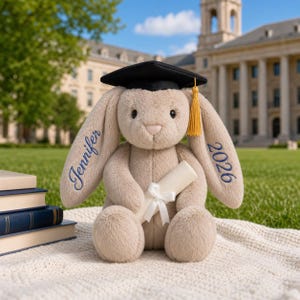 May include: A plush bunny wearing a black graduation cap with a gold tassel. The bunny holds a rolled diploma tied with a white ribbon. The name "Jennifer" and the year "2026" are embroidered in blue on the bunny's ears. The bunny is sitting on a white blanket.