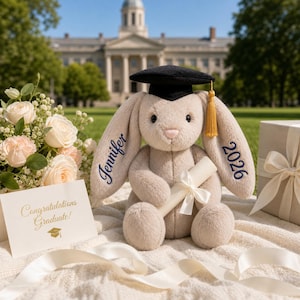 May include: A plush bunny wearing a graduation cap and holding a diploma. The bunny's ears are embroidered with "Jennifer" and "2026". A card reads "Congratulations Graduate!" with a graduation cap icon. A bouquet of flowers and a gift box are also present.