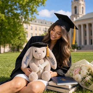May include: A plush bunny wearing a graduation cap and holding a diploma. The bunny has "Jennifer" embroidered on one ear and "2026" on the other. The bunny is tan with a black graduation cap and gold tassel. A bouquet of flowers and books are nearby.
