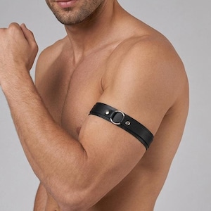 May include: A black leather arm band featuring a central silver-toned metal O-ring and two silver-toned metal rivets. The band is constructed from a smooth, dark material and is designed to be worn around the upper arm as a decorative accessory.