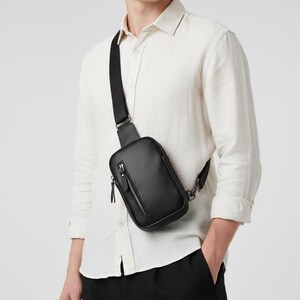 May include: Black crossbody bag with a textured surface and a zippered front pocket. The bag has a black adjustable strap and a silver-tone metal clasp. The bag is worn over the shoulder.