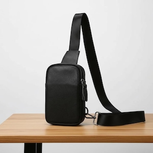 May include: Black crossbody bag with a textured leather-like finish. The bag features a rectangular shape with a zippered main compartment and a smaller front pocket. It has a long, adjustable black strap and a metal clasp.