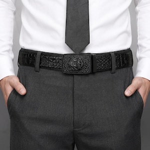 Op de afbeelding: Een zwarte riem met een textuur van krokodillenleer en een rechthoekige gesp met een reliëf van een dierenkop. De riem wordt gedragen over een donkergrijze broek met een wit overhemd en een effen zwarte das.