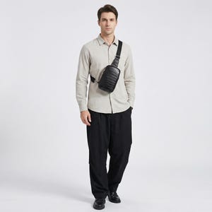May include: A black leather crossbody bag with a textured pattern is worn across the chest. The bag has a rectangular shape and a black adjustable strap. The person is wearing a light-colored button-down shirt and black pants.