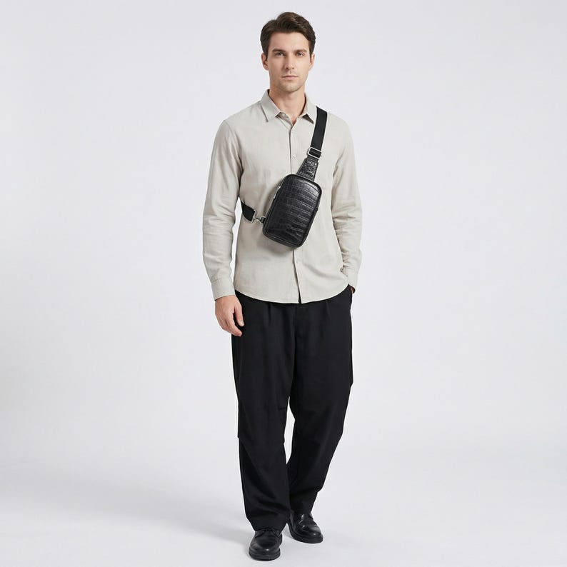 May include: A black leather crossbody bag with a textured pattern is worn across the chest. The bag has a rectangular shape and a black adjustable strap. The person is wearing a light-colored button-down shirt and black pants.