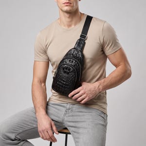 May include: A black sling bag with a textured crocodile skin pattern, featuring a main zippered compartment and a smaller front zippered pocket. The bag has an adjustable black fabric strap with a silver-toned metal buckle for secure closure.