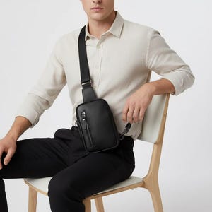 May include: A black leather crossbody bag with a zippered front pocket and adjustable strap. The bag is worn across the body. The bag is approximately 9 inches tall and 6 inches wide.