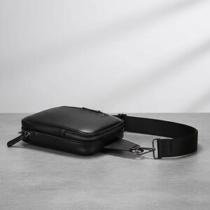 May include: A black leather crossbody bag with a rectangular shape and a zippered main compartment. The bag features a long, adjustable black strap and silver-toned hardware. The bag is designed to be worn across the body.