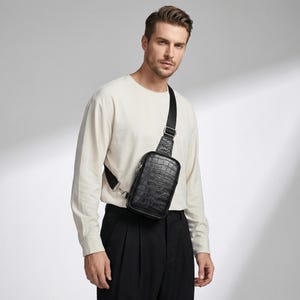 May include: Black crossbody bag with a textured, crocodile-like pattern. The bag has a rectangular shape with a zippered closure and an adjustable black strap. The bag is worn across the body.