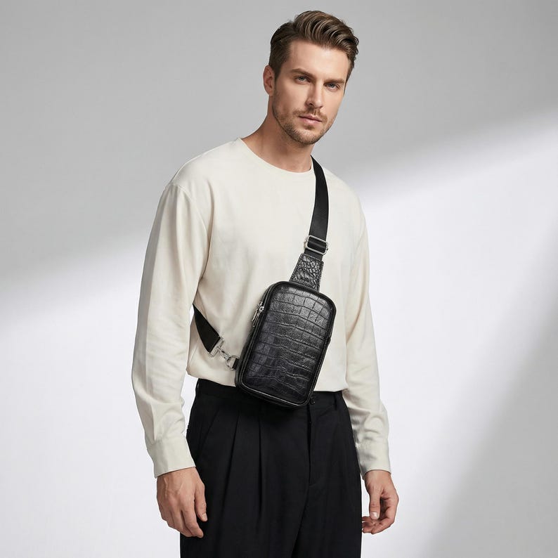 May include: Black crossbody bag with a textured, crocodile-like pattern. The bag has a rectangular shape with a zippered closure and an adjustable black strap. The bag is worn across the body.