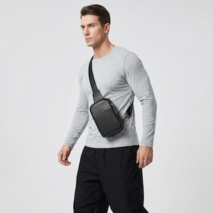 May include: A black crossbody bag with a rectangular shape and a zippered front pocket. The bag is worn over the shoulder with a black strap. The person in the image is wearing a gray long-sleeve shirt and black pants.